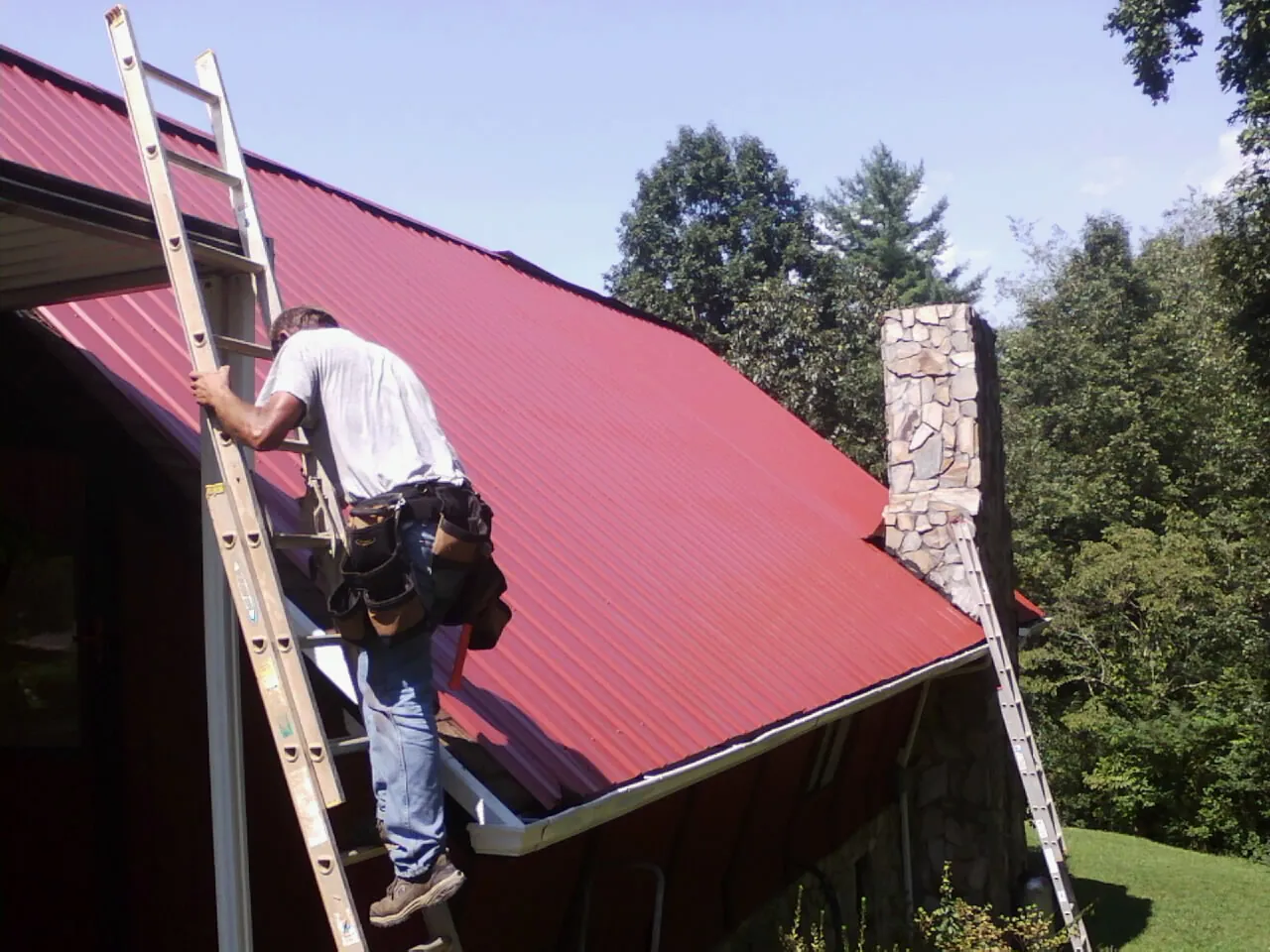 Roofer on a ladder beside a red metal roof and stone chimney — Soffit & Fascia Repair by Roofing Brothers in Waddell